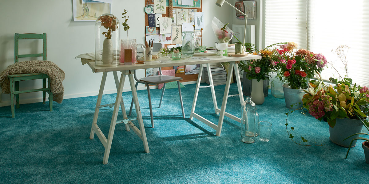Ein blauer Teppichboden in einem Zimmer mit einem Tisch und Blumen. Boden Pelzer -Bodenbeläge, Vinylbelag, Parkett - Moselstr. 14 - 41464 Neuss